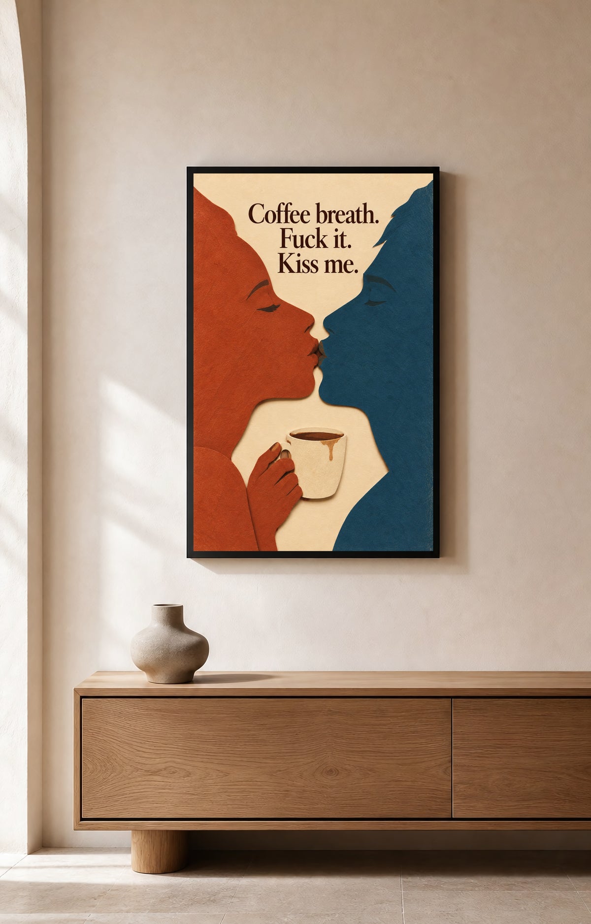 Coffee Breath shown as framed wall art in a room. Burnt-orange and deep-teal profiles kiss above a cream coffee cup; the artwork reads Coffee breath. Fuck it. Kiss me.